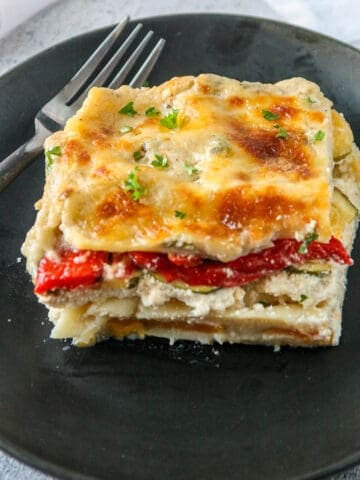 A slice of vegetable lasagna with white sauce on a black plate.