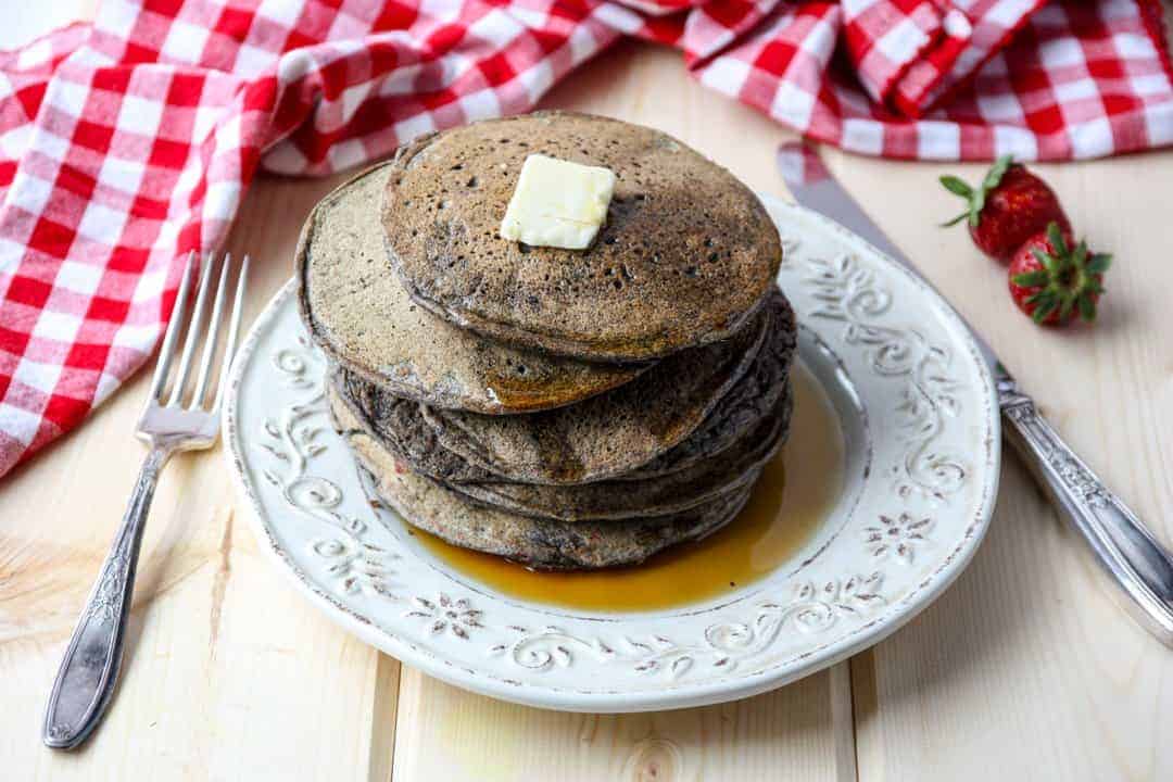 Stack of buckwheat pancakes on an embossed white plate with a pat of butter on top and maple syrup