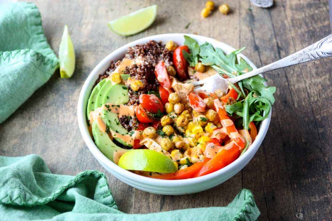 Fresh vegetables and grains in white bowl with fork on wooden table.