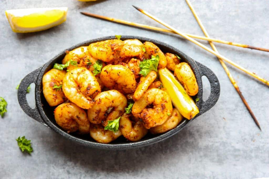 A black dish filled with seasoned and cooked shrimp garnished with fresh parsley. The dish is accompanied by lemon wedges and three wooden skewers are placed in the background on a gray surface.