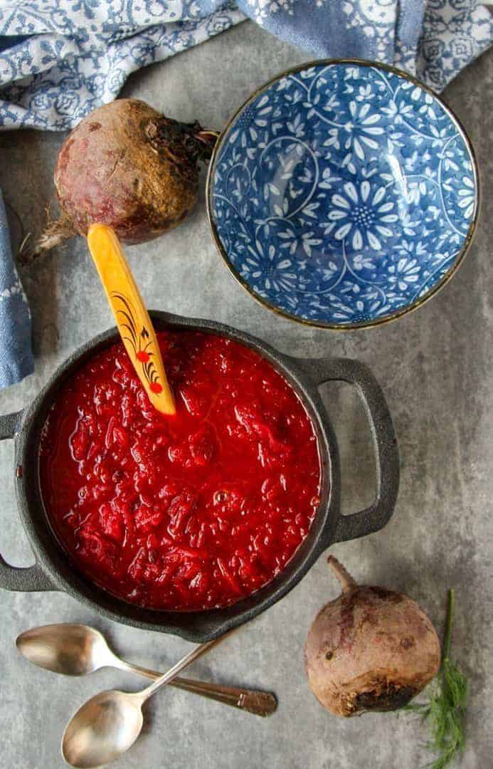 top shot of red beet soup in black pot with wooden spoon next to blue and white floral bowl and two silver spoons