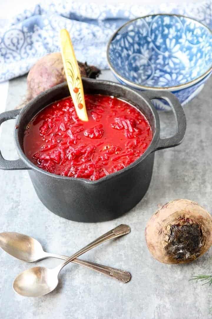 red beet soup in black pot with wooden spoon next to blue and white floral bowl and two silver spoons