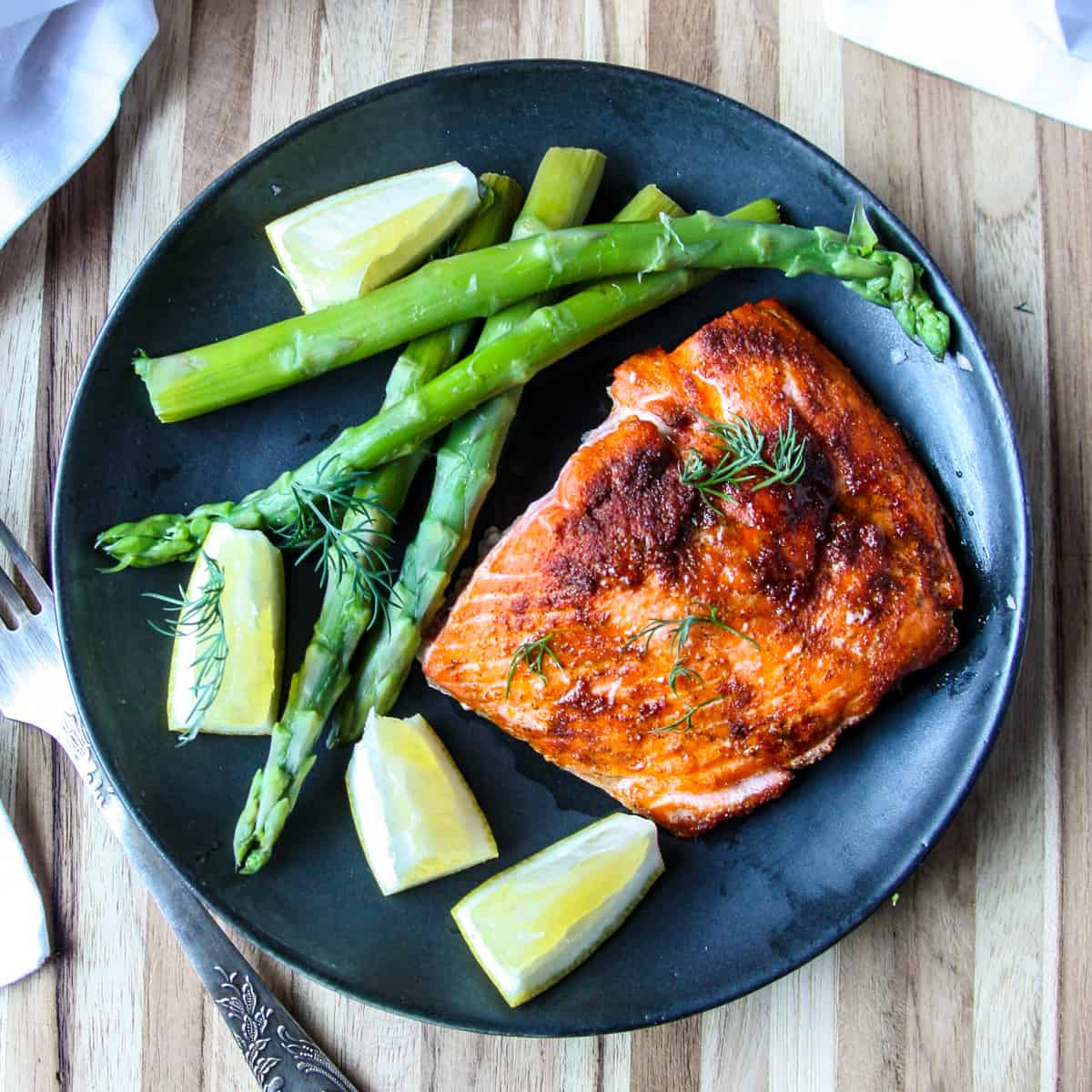 An air fryer cooked fillet of salmon on a black plate with asparagus and lemon wedges.