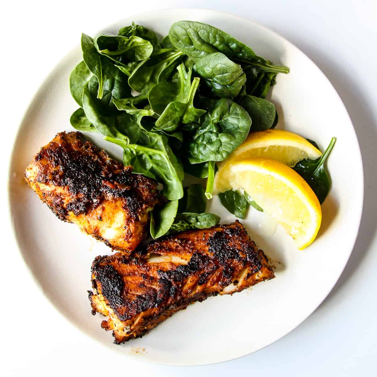 2 blackened cod loin pieces on a white plate with lemon wedges and salad greens.