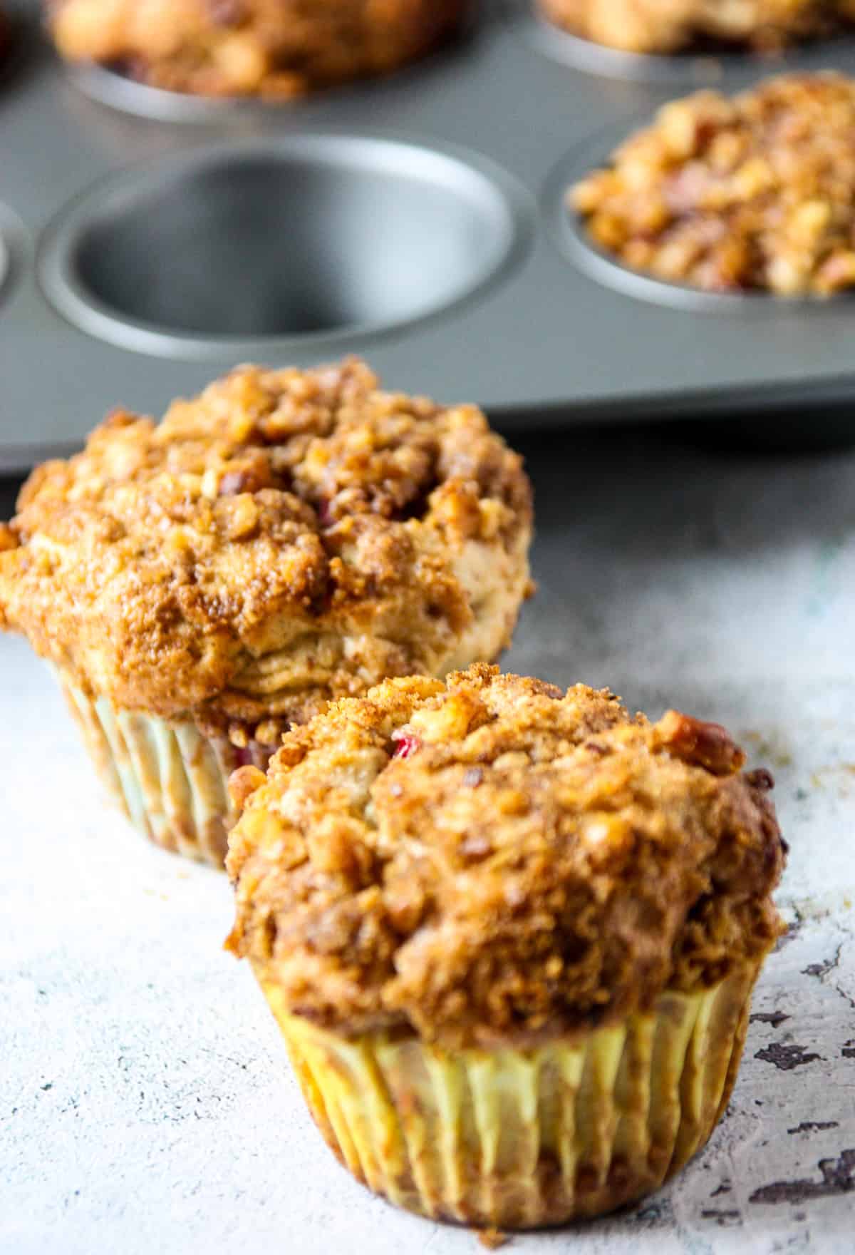 Two strawberry rhubarb muffins with a golden streusel topping on a stone counter top.