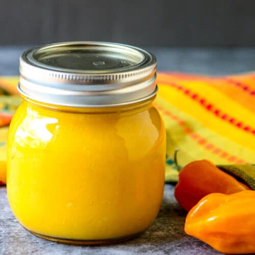 A glass jar of bright yellow mango habanero sauce, next to two habanero peppers.