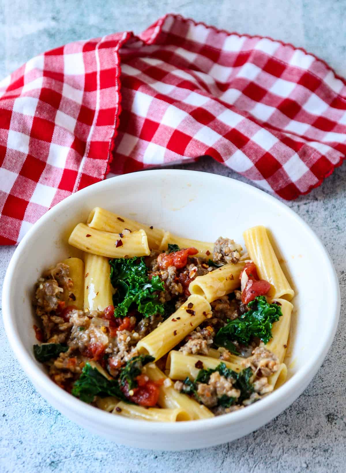 Sausage and kale pasta in a white bowl next to a red and white cloth napkin.